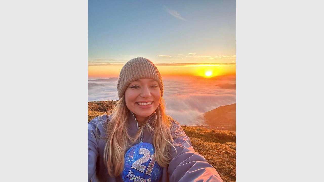 Honor is wearing a hat with her blonde hair down and a big badge saying '21 today' on the front of her jacket. She's holding the camera out to take a selfie and she's smiling while stood with mountains and a bright sunrise behind her. 