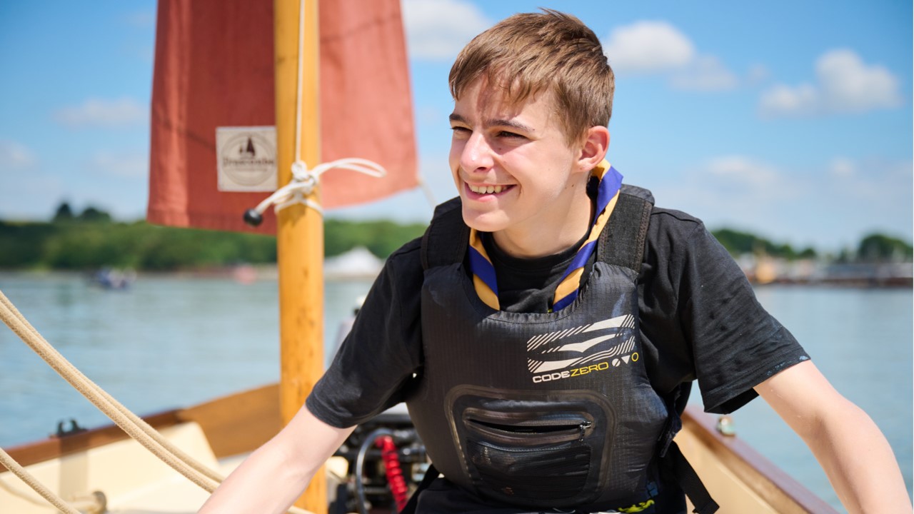 A young person, wearing a life jacket and a yellow and blue necker, smiles while sat on a sailboat.