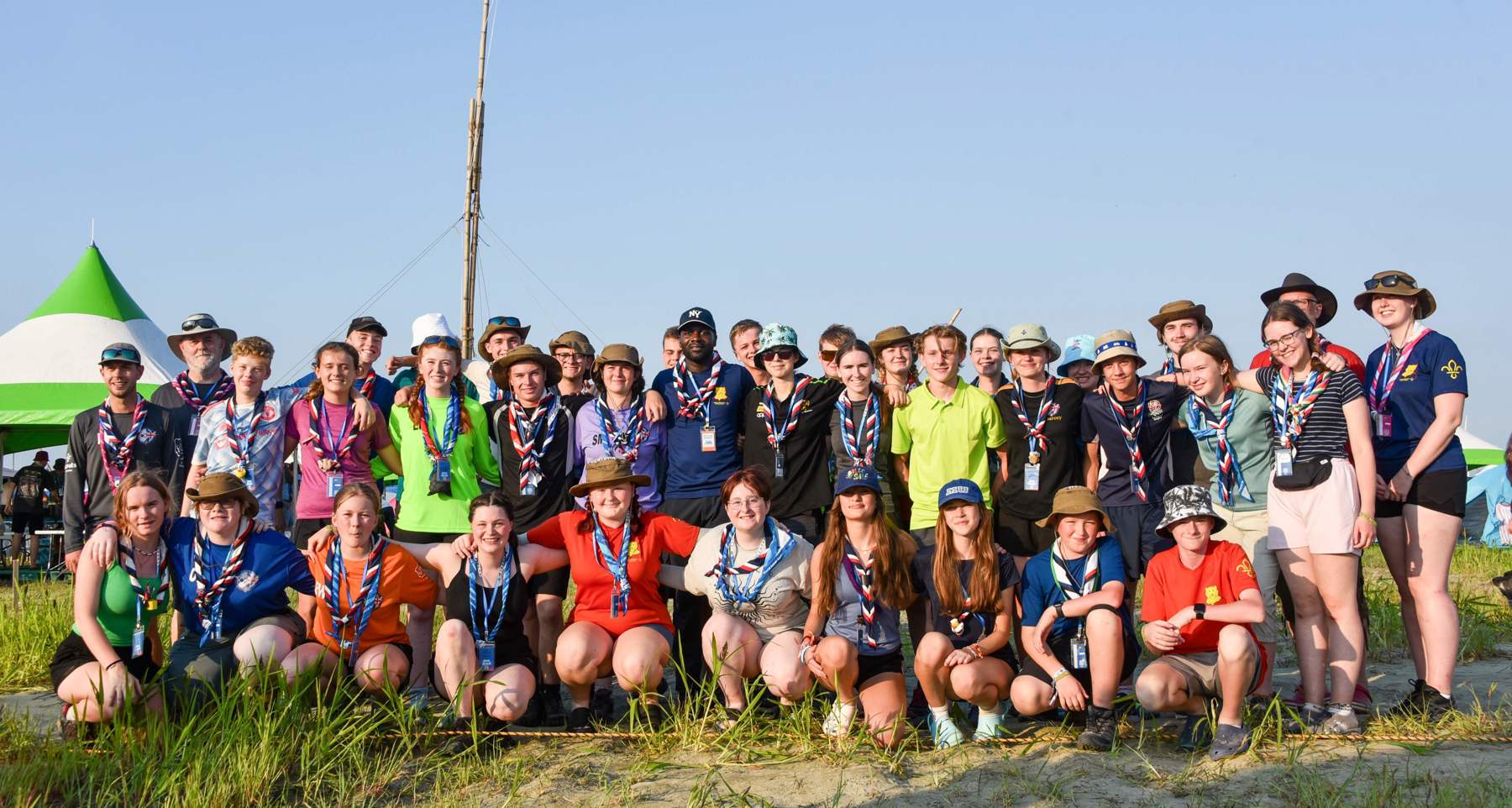 A large group of Scouts pose together outdoors on a sunny day, many wearing colorful shirts, neckerchiefs, and hats. They are smiling and standing or kneeling on sandy ground with some grass. Green and white tents are visible in the background under a clear blue sky at the World Scout Jamboree in Korea 2023.
