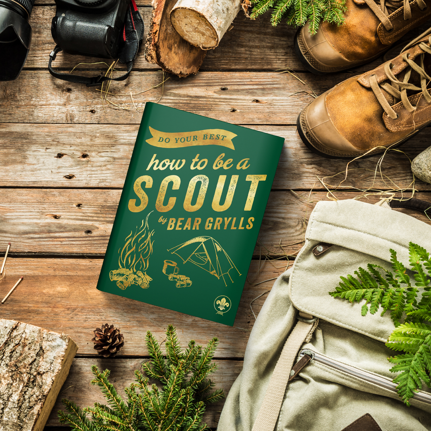 The image shows Bear Grylls' latest book, Do Your Best: How to be a Scout. The book is laid on top of wooden planks and is surrounded by different outdoor items, including brown boots, a backpack, leaves, and a pine cone. 