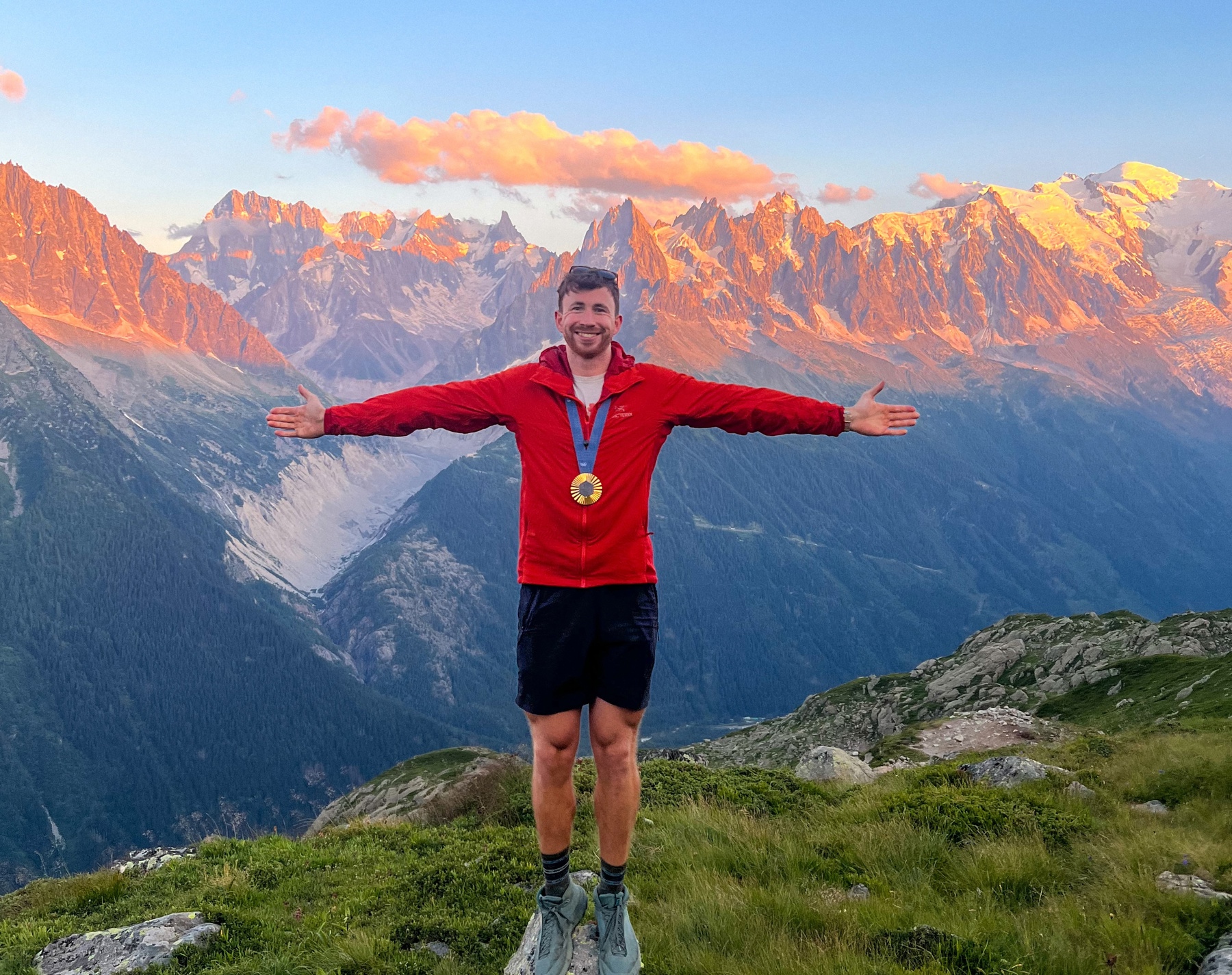The image shows Morgan standing on the top of a grassy mountain, wearing a red coat, navy shorts and blue trainers. He's holding his arms out horizontally either side and is smiling at the camera. There are clouds and mountains in the background, with a blue sky and a faint orange hue across the mountains as the sun sets.