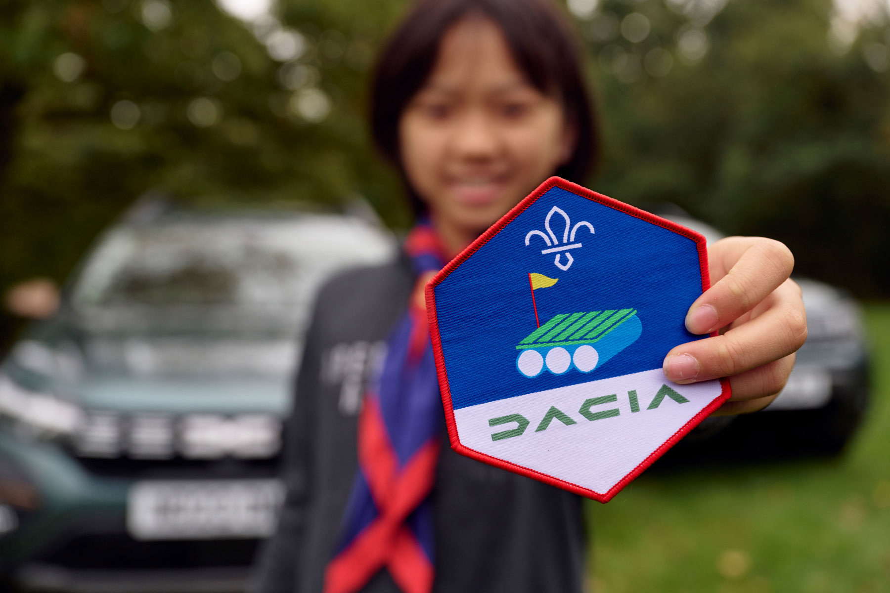 The image shows a Scout wearing a purple and red necker and stood in front of two cars. The Scout and the cars are blurred in the background, with the focus on the Dacia badge, which the Scout is holding out right in front of the camera.