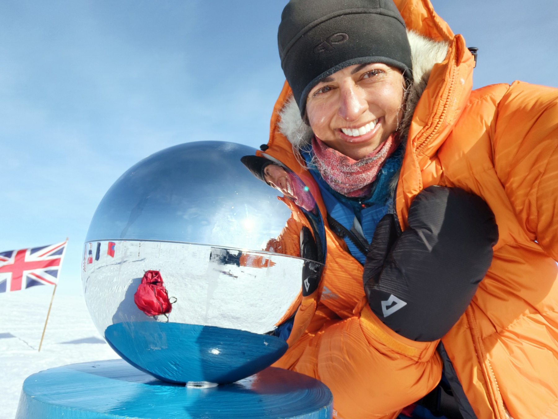 The image shows Preet Chandi MBE wearing an orange coat, black gloves and a hat, crouching down to take a selfie while standing on snow. There's a Union Jack flag in the ground behind her and a mirrored statue next to her on the left.