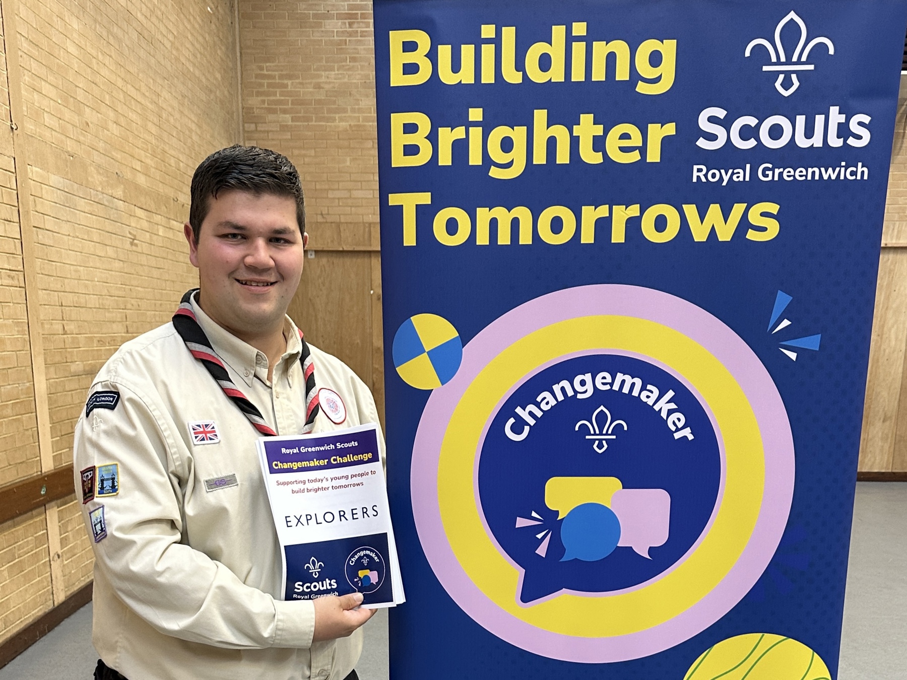 Joseph is stood inside of a hall to the left holding the Changemaker Challenge pack that he created. He's wearing his Scout volunteer uniform with a necker and he's smiling at the camera. To the right of him is a standing banner that says 'Building Brighter Tomorrows' with the Changemaker badge logo underneath. There's a white Scouts Royal Greenwich logo in the top left corner.