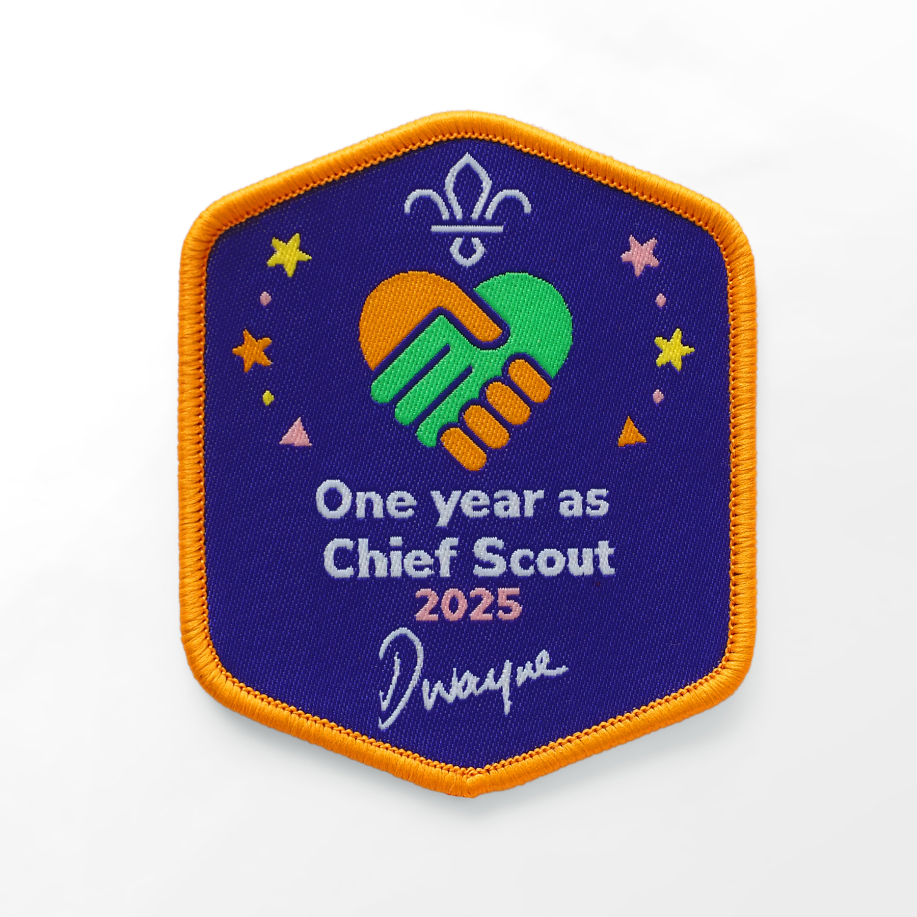 The image shows the One year as Chief Scout blanket badge. It's got a purple background with a orange edge. There's a white fleur-de-lis at the top in the middle, and a green and orange heart and hands image beneath it with stars. The text says 'One year as Chief Scout 2025' with Dwayne Field's signature underneath.