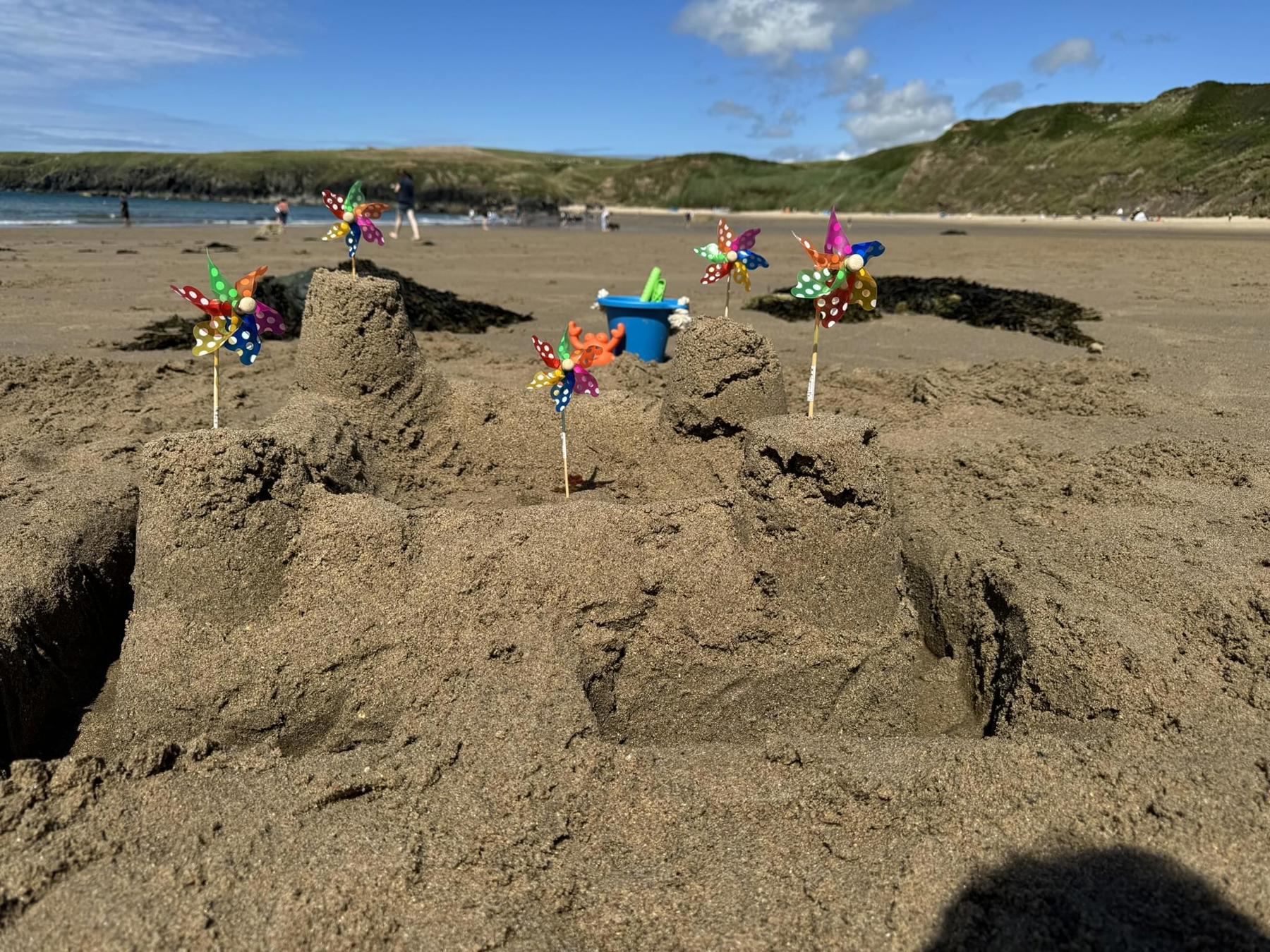 A sandcastle on a beach with a blue sky in the background. The sandcastle has four turrets in each corner, with walls joining them and a windmill in each. There is a moat around it with a bridge. 