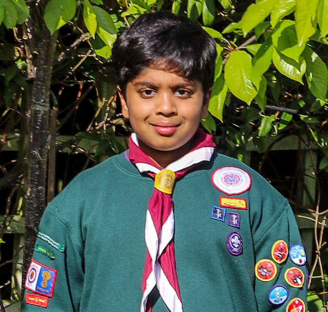 Aneeshwar, wearing a teal jumper with lots of badges and a white and red necker, looks at the camera while smiling.