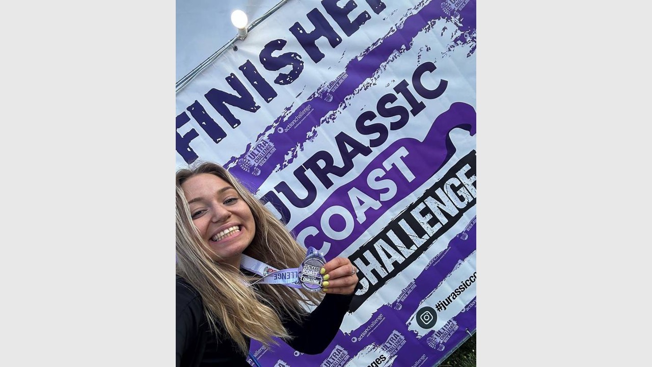 Honor is taking a selfie in front of the finishing banner for the Jurassic Coast Challenge. She's smiling at the camera while holding her medal up. 