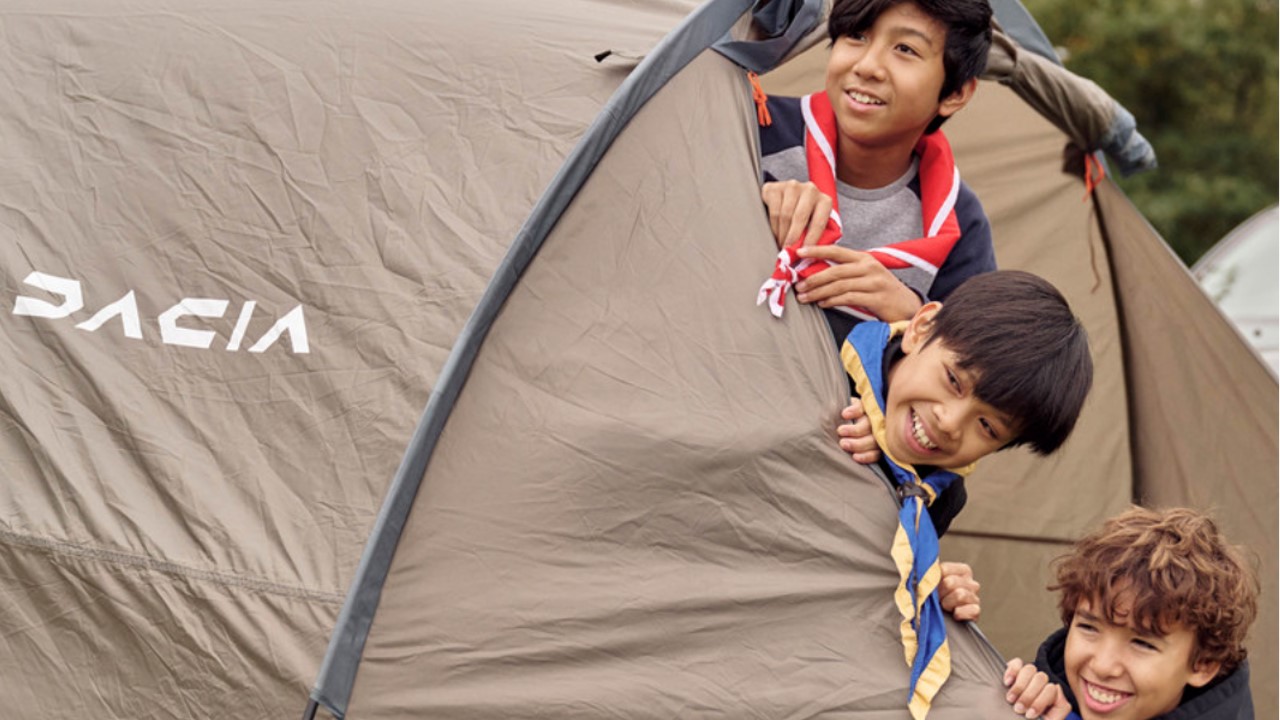 Three young people smile while sticking their heads out of a tent that says 'Dacia' on the side.