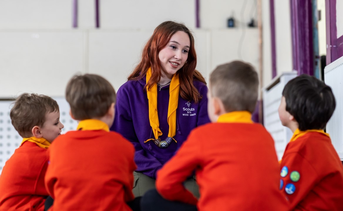 A volunteer, wearing a purple jumper and orange necker, smiles while talking to a group of Squirrels.