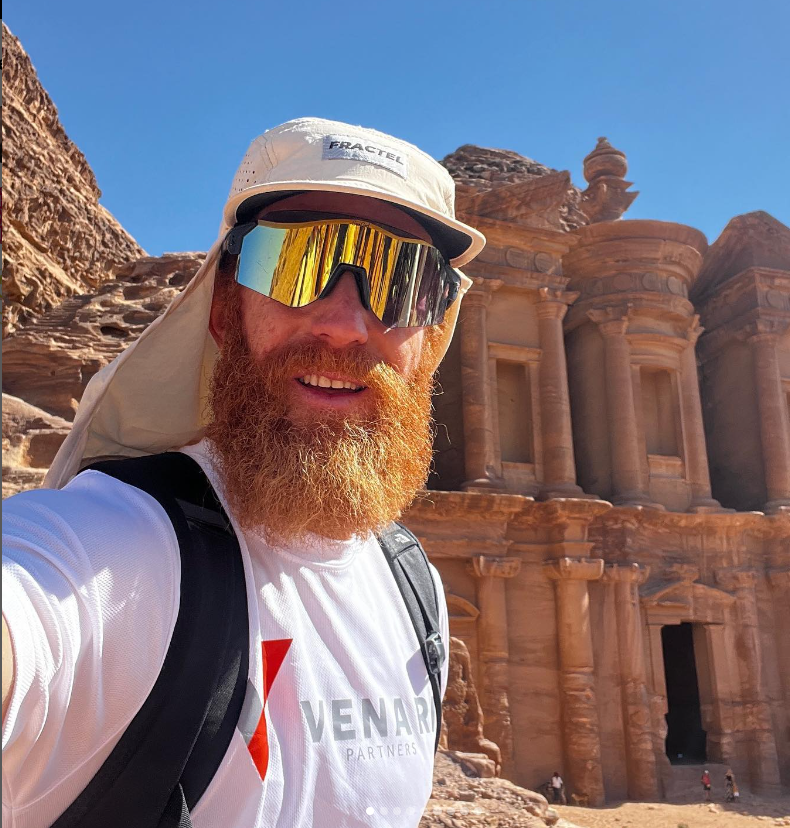 Sean's wearing goggles and a hat as he stands in the sun in Petra. He's stood in front of an old building and is holding his camera with his left hand as he takes a selfie.