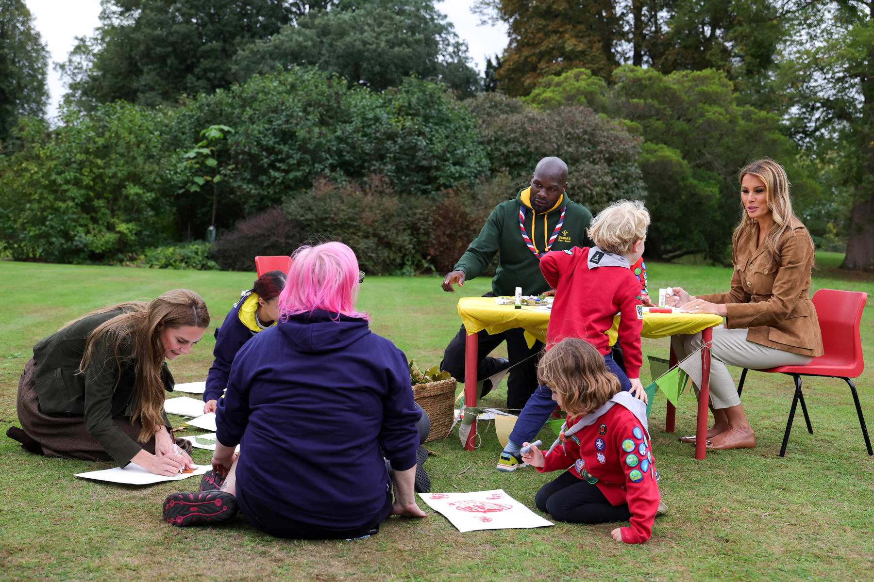 The image shows HRH The Princess of Wales, Chief Scout Dwayne Fields, First Lady of the United States, a Scout volunteer and two Squirrels on grass doing drawing. There's a table set up and paper on the floor for crafts.