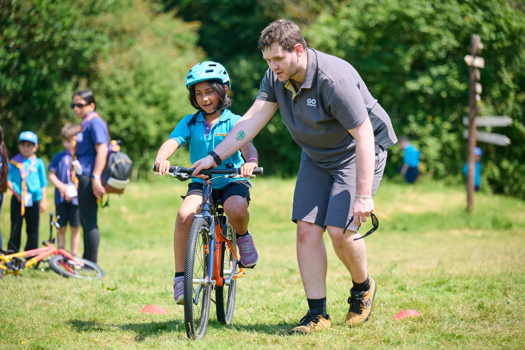 A Beaver is wearing a blue Beavers polo shirt and a blue helmet and is riding a bike on grass. To the right of her is a GO Outdoors volunteer holding onto her handlebars with his right hand. There are two red cones on the grass just behind them, and there are two other Scouts and a volunteer in the background to the left of them.