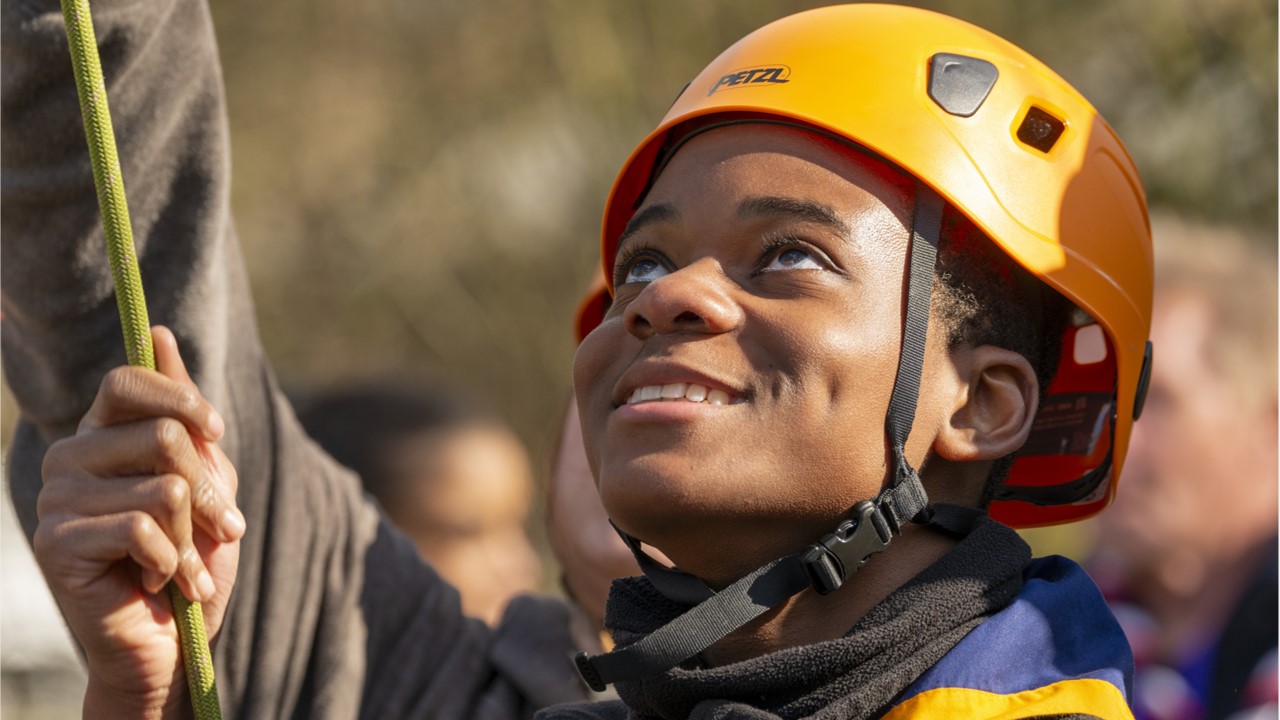 A young person, wearing an orange helmet, holds onto a rope while looking up and smiling.