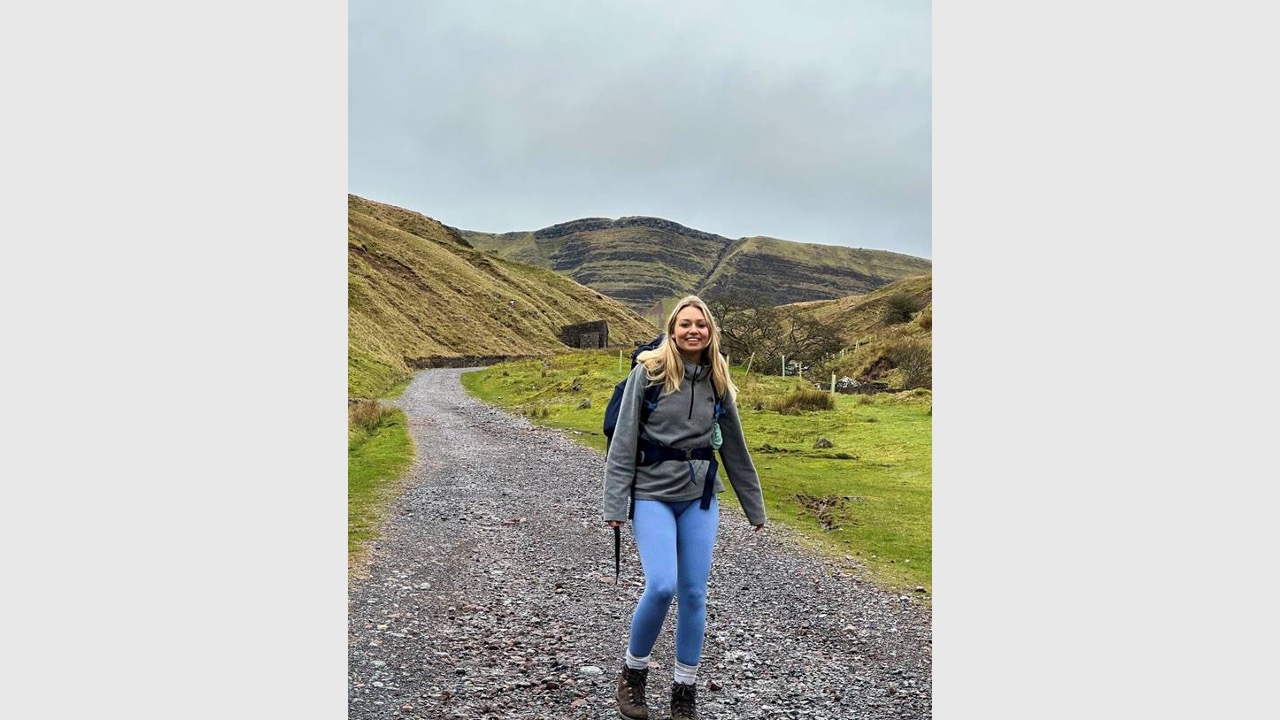 Honor is wearing a grey jumper, leggings, hiking boots and a backpack as she stands on a gravel path smiling at the camera. Behind her is green grass and mountains, and a grey cloudy sky.