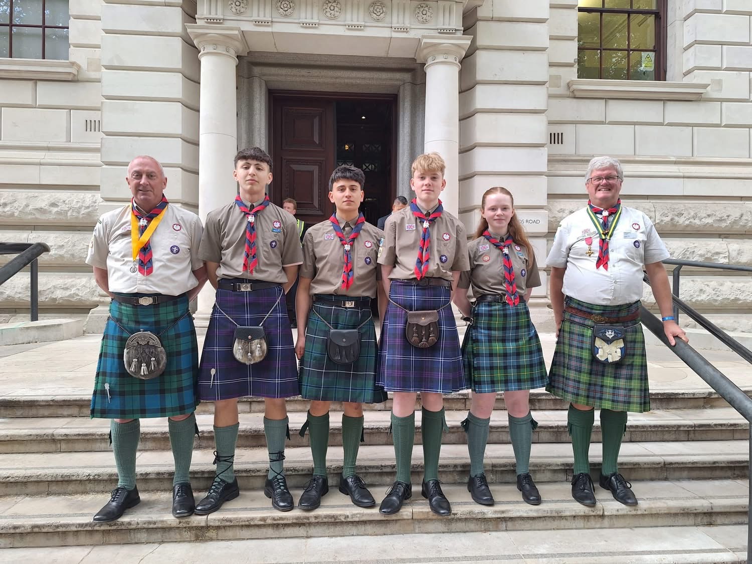 The image shows two volunteers and four Scouts at the procession as part of the VE80 celebrations. They're all wearing kilts, shirts and neckers and are stood with their hands behind the backs on concrete steps.