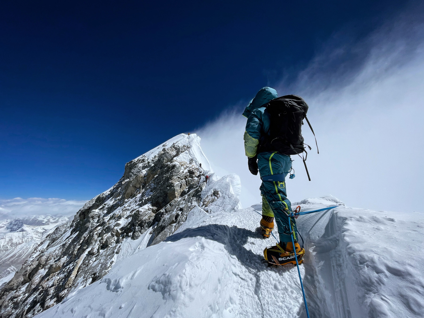 Kirsty has her back to us and is close to the top of Mount Everest. She has a black rucksack on her back, snow boots on, a rope attached to her, her hood up and gloves on. She's looking to the highest point of Everest with clouds to her right and a bright blue sky.