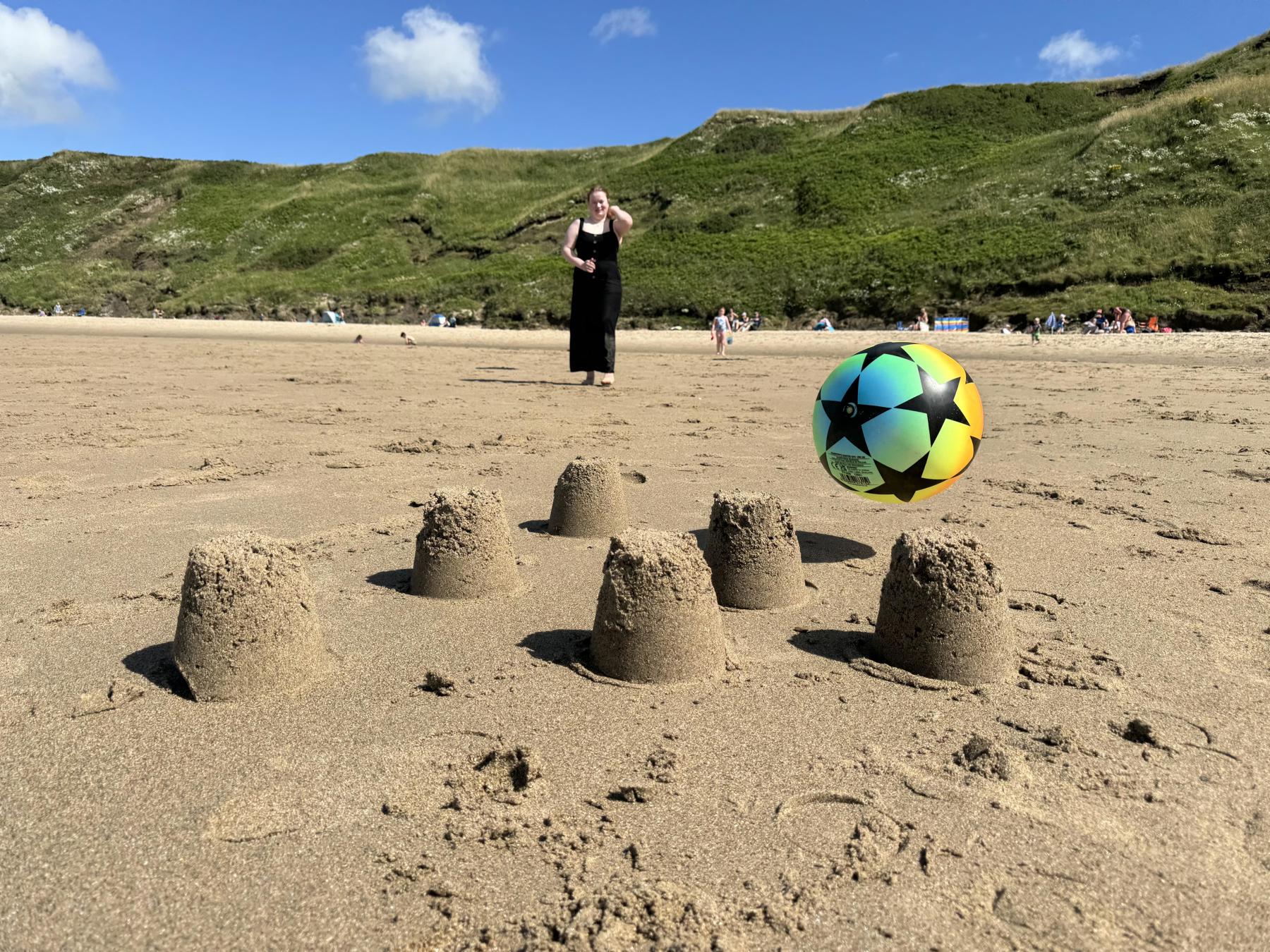 Six sandcastles are on the beach and someone is throwing a ball towards them. There is green grass and blue skies behind the beach 