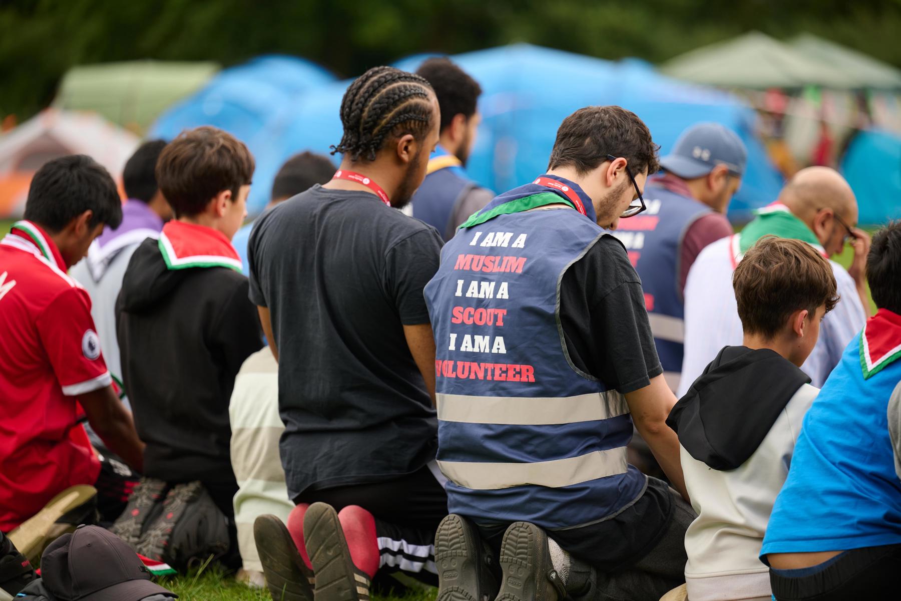A group of young people and volunteers kneeling down. One is wearing a vest that says "I am a Muslim, I am a Scout, I am a volunteer".