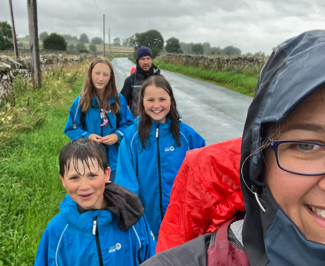 The image shows the Great Woggle JOGLE team walking by a road on a grass verge wearing raincoats. There are grey clouds in the sky and it's raining.