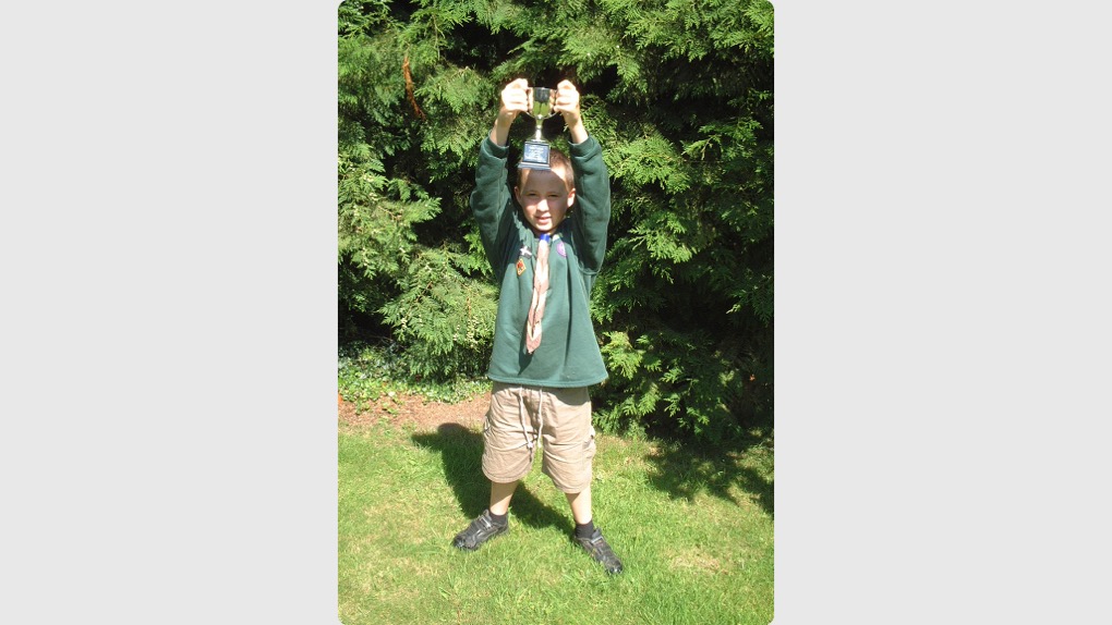 The image shows Morgan standing outside on grass in front of a hedge on a sunny day. He's wearing his Cubs uniform and a cap while holding a trophy for winning Cub Scout of the Year in both hands above his head.