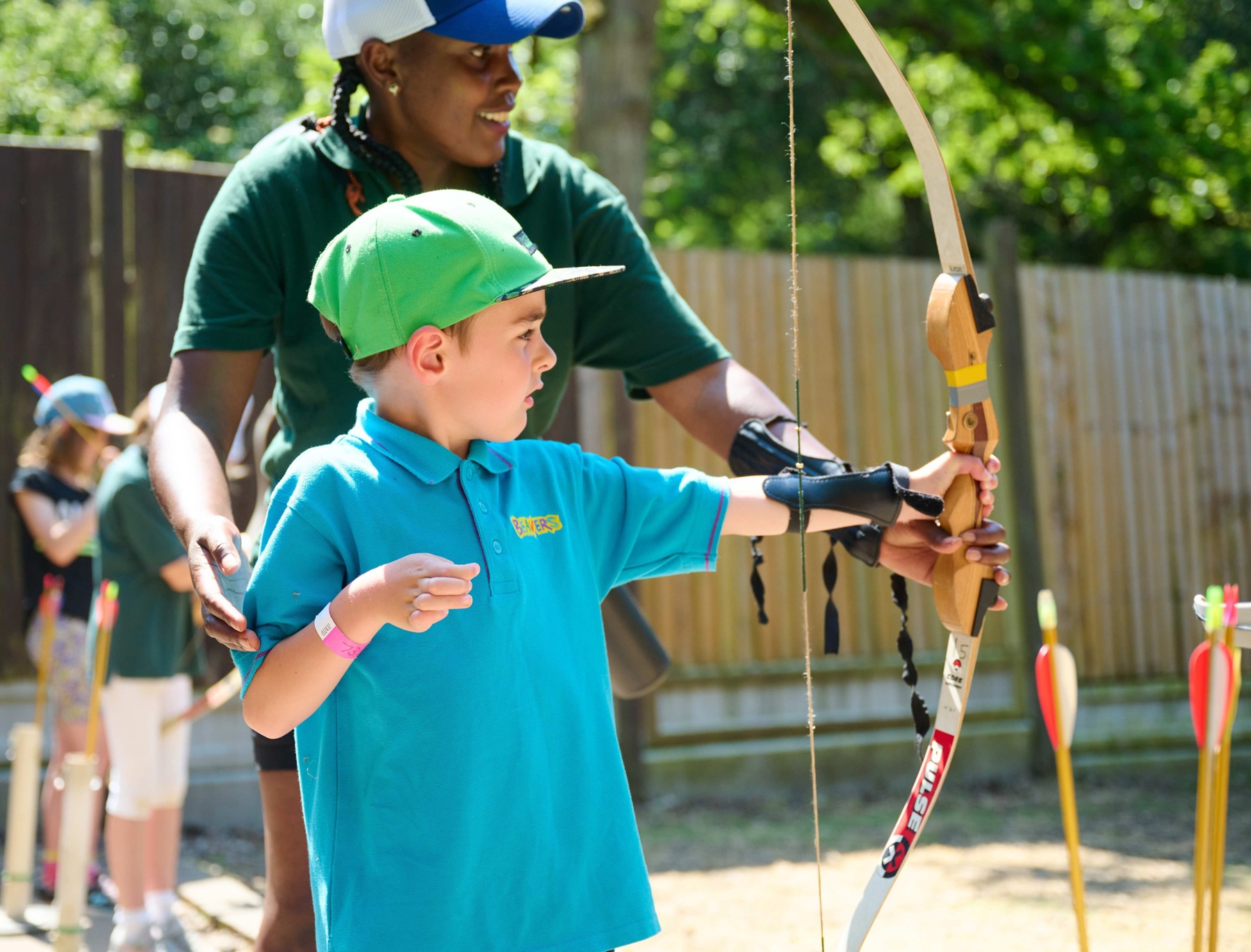 The image shows a Beaver wearing a Beaver blue polo shirt and a green hat, facing to their left. They're holding an archery bow and there's a volunteer in a dark green polo and cap stood behind them to help. The sun is shining and they're outside, and there are other Beavers/Cubs in the background by a wooden fence.