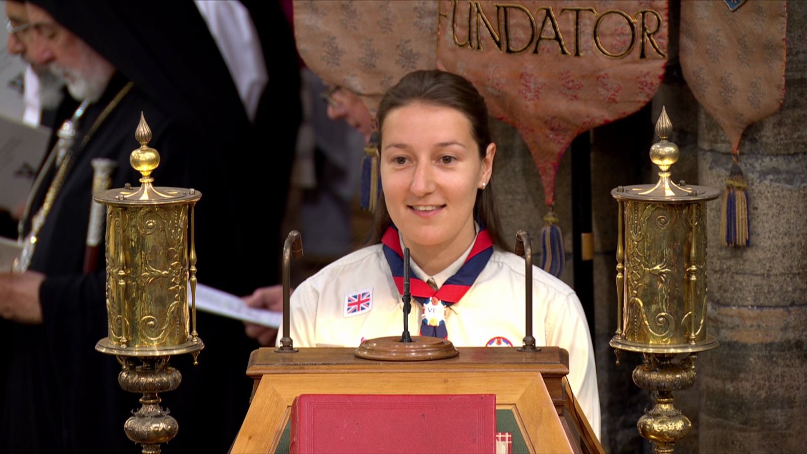 The image shows Abbie McFarlane, King's Scout, standing by a podium in Westminster Abbey for the Service of Thanksgiving as part of the VE80 celebrations. She's wearing her uniform and necker with a VE Day 80 woggle.