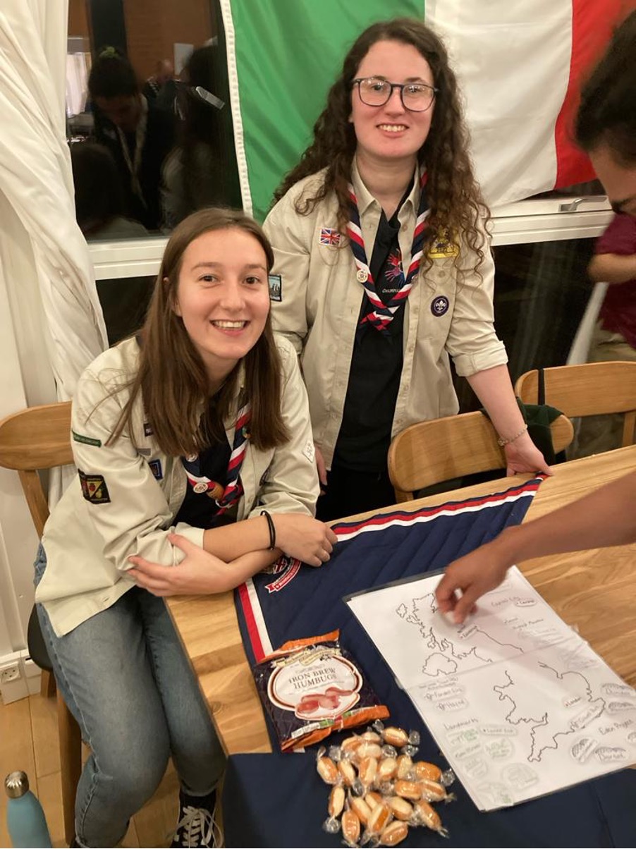 Alice is sat on a chair wearing a beige Scout blouse and red, navy and white necker, sitting next to her fellow UK Rep Pool member, Abbie. Abbie is stood by a wooden chair and both are smiling at the camera, with their hands on the wooden table in front of them. There's a flag and window behind them. 