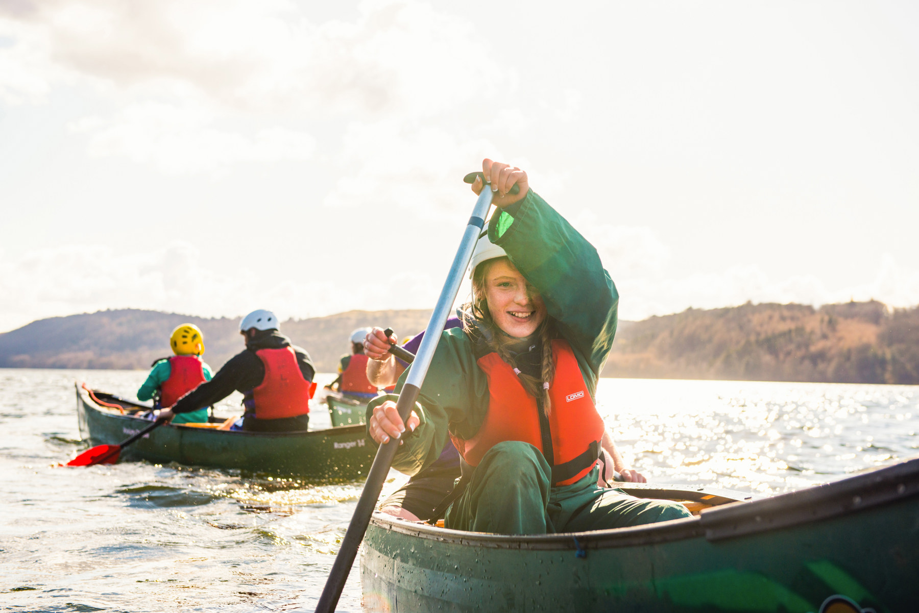A young person paddling a canoe on open water smiling to the camera. There are other young people in canoes on the water too.