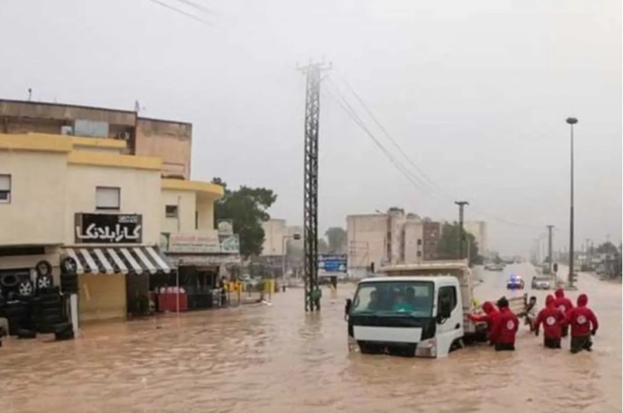 The image shows flooding in Libya. To the left there is a building, a telephone pole in the middle, and to the right workers next to a truck that's half under the water.