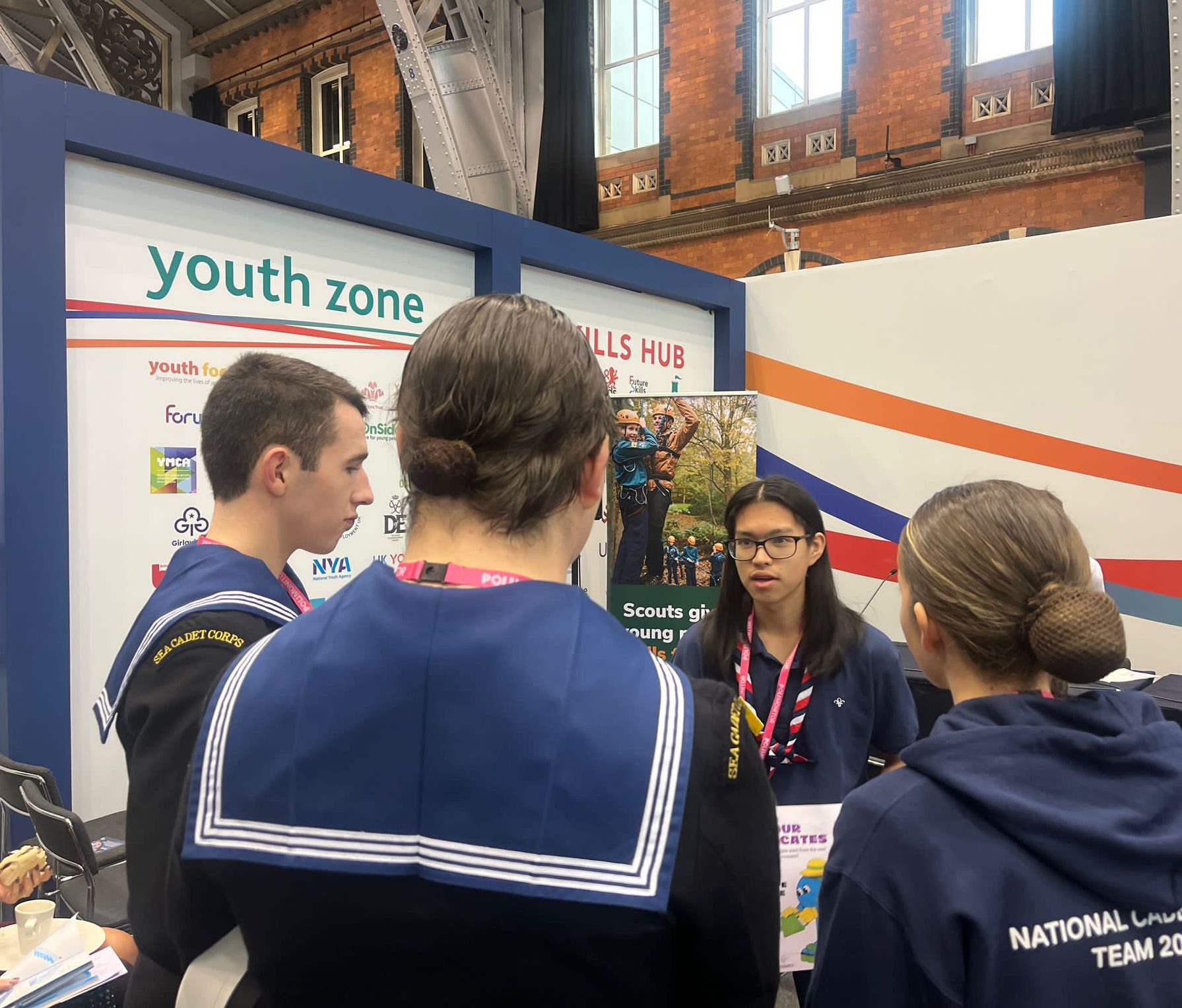 Three Sea Cadets are stood with their backs to the camera wearing navy clothes. They are speaking to Dan, a Scouts Youth Advocate, who is facing the camera and wearing a Scouts necker and navy polo shirt. To the left. is a poster that says 'Youth zone'.