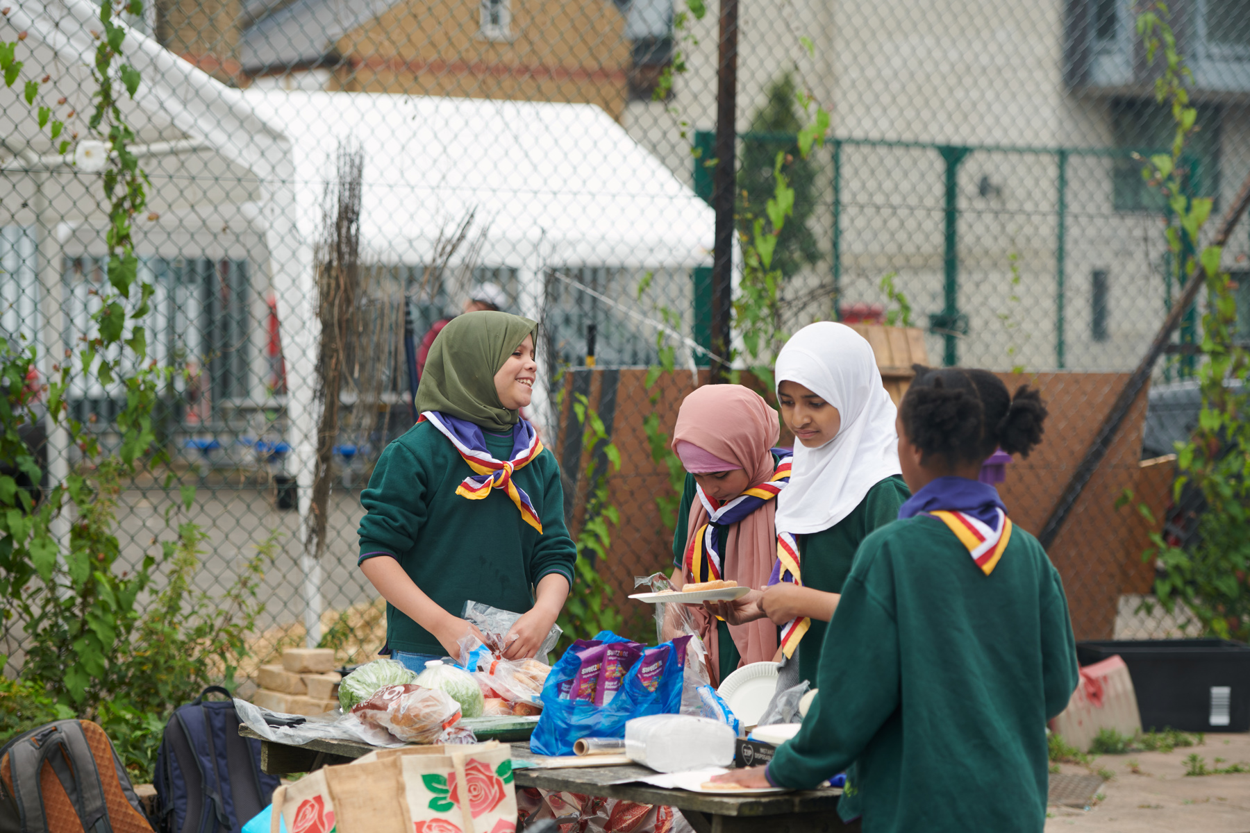 A group of young Muslim Scouts are wearing their uniform and neckers. One Cub on the left is looking at the others to the right and smiling, with the other three looking down at the food on a table outside. They're stood in front of a fence and there's bread, carrier bags, lettuce and other packets of food on the table. Around the table is a shopping bag and a few backpacks.