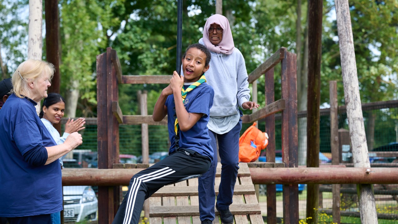 A young person, wearing a blue t-shirt and yellow and green necker, goes down a zipline while volunteers watch on.