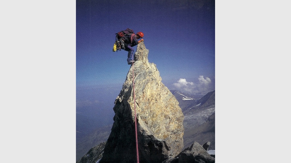 The image shows Karen Darke climbing a narrow mountainside in the French Alps when she was 21. The image shows her clinging onto the top of the mountain with a rope attached to her and she's wearing a red helmet and backpack. There are mountains, clouds and blue sky in the background.