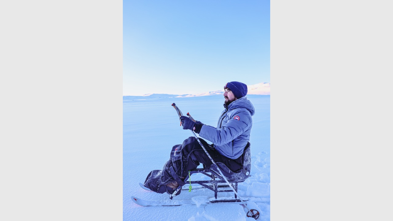 Darren is sat down on snow, wearing a winter outdoor gear. He's holding poles and the camera is taking the photo side on.