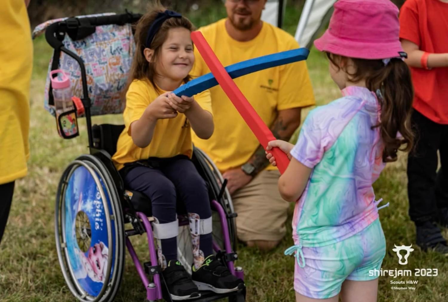 The image shows Sophie at ShireJam 2023. She's in her wheelchair on grass opposite another Scout. She's holding a blue foam sword up to the other Scout's red one. There's a volunteer in a yellow top crouching down behind her.