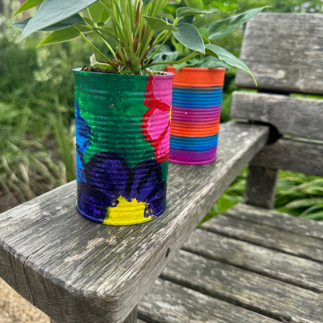 Two tin cans with plants in. They've been painted brightly, one with flowers and one with stripes, and are resting on a park bench arm.