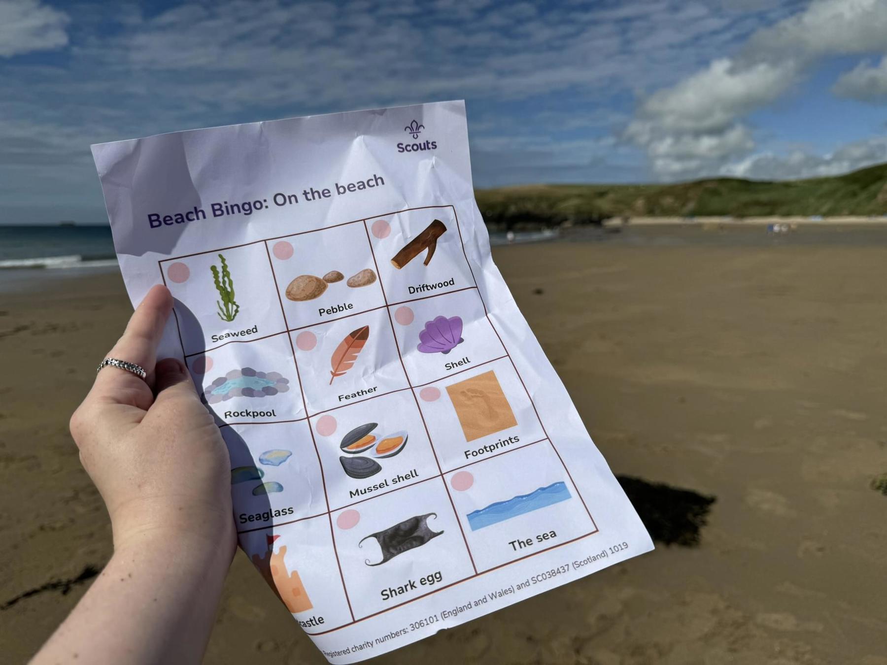 A hand is holding up a sheet of paper with different objects to tick off that you might find on a beach, like a bingo card. In the background there is sand and sky.