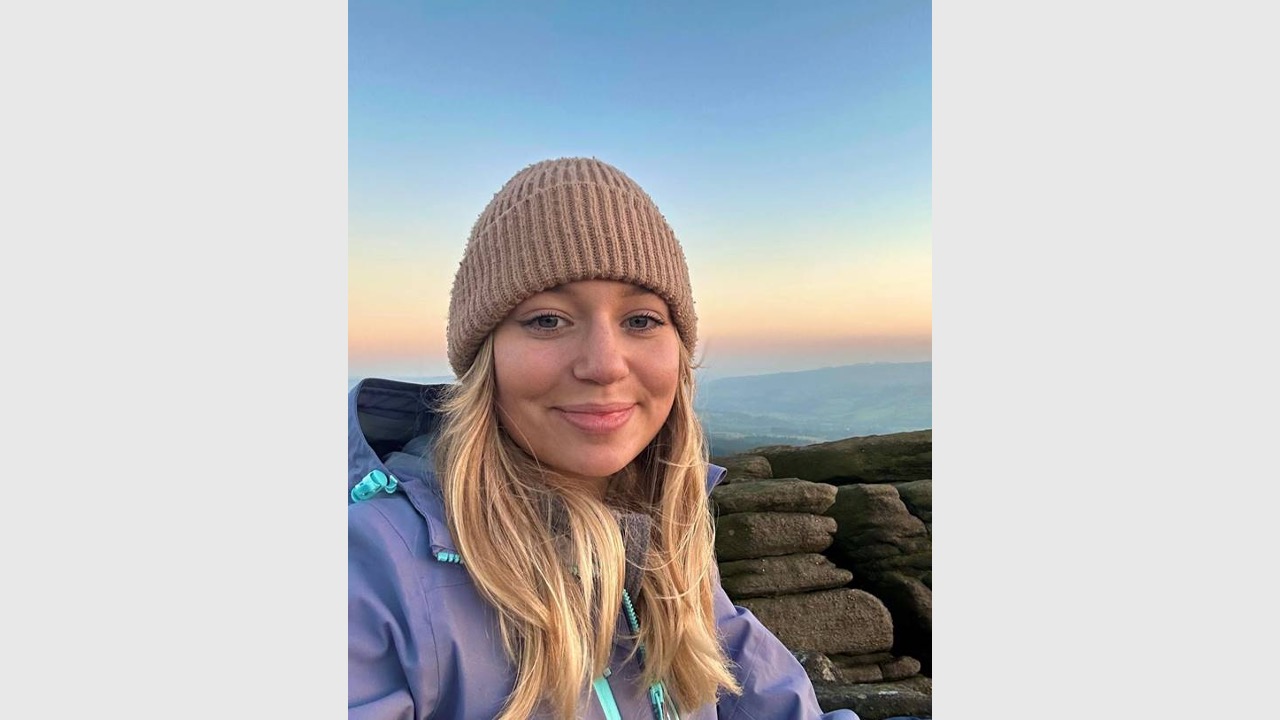 Honor is taking a selfie wearing a hat and jacket. She's stood in front of a sunrise with the ocean and rocks behind her. 