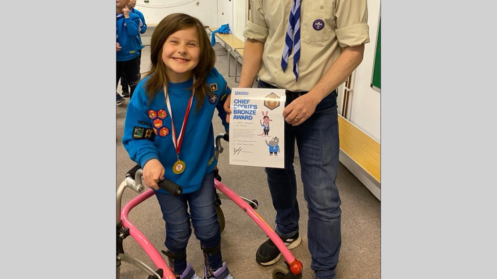 The image shows Sophie standing in her Beaver uniform with badges on her blue jumper and a medal around her neck. She's smiling at the camera and there's a volunteer in uniform to her right who's holding her Chief Scout's Bronze Award certificate.