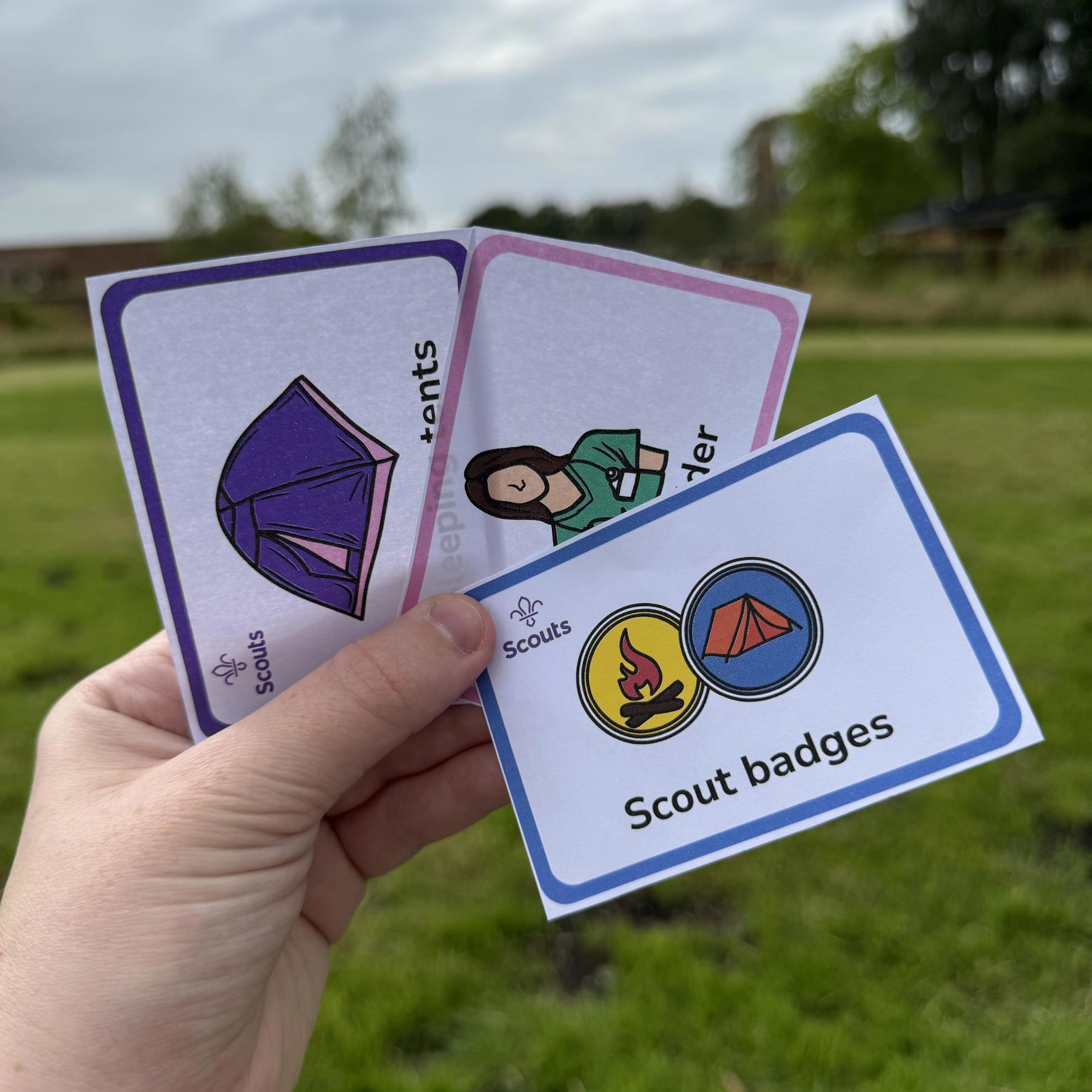 Image of someone holding some Scout themed cards in their hand, with a field and trees behind them. There's three paper cards. One has a purple border, with a picture of a purple tent. One has a pink border and picture of a first aider in green uniform. The last one has a blue border with a picture of two Scout badges on - one badge has a picture of a campfire and the other badge has a picture of a tent.