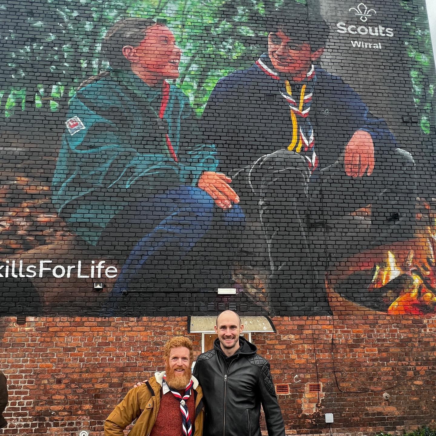 In the image, Sean Conway stands with artist Paul Curtis in front of the Wirral Scouts mural. Sean's wearing a red white and navy necker and is stood next to Paul on his right hand side. Both are smiling at the camera.