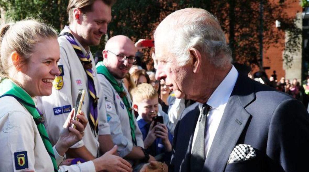 King Charles III greets a line of smiling Scouts wearing beige shirts and neckers.