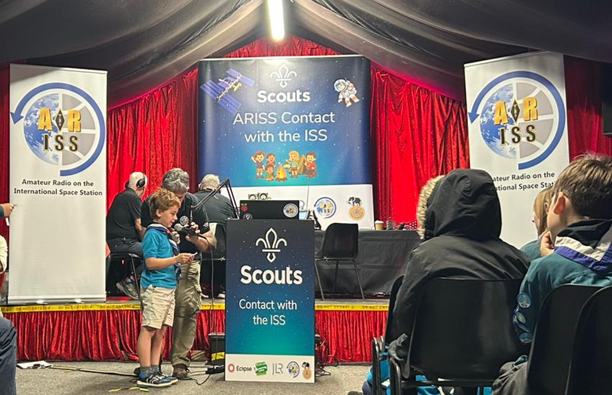 A young Scout speaks into a microphone while holding a piece of paper at an event called “Scouts ARISS Contact with the ISS.” Behind him, adults operate radio equipment on a stage decorated with red curtains. Banners on either side display the ARISS (Amateur Radio on the International Space Station) logo. Audience members, including other Scouts, watch the interaction.
