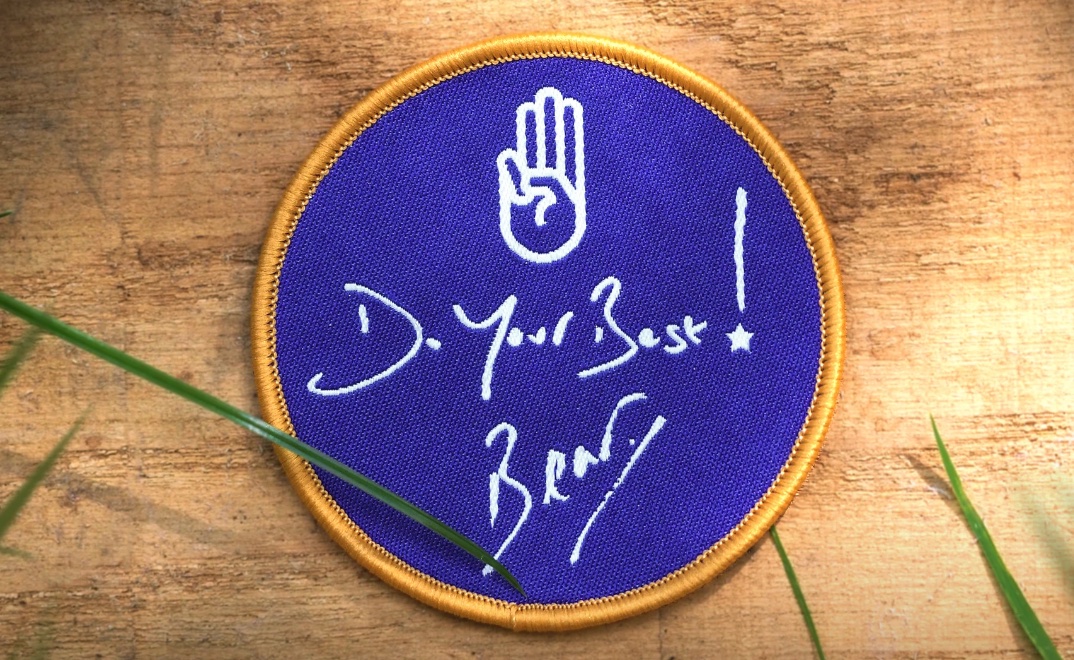 The image shows a navy badge with a gold border that says 'Do Your Best' with Bear's signature in white underneath. The badge is on a wooden background and there are a few blades of grass surrounding it.