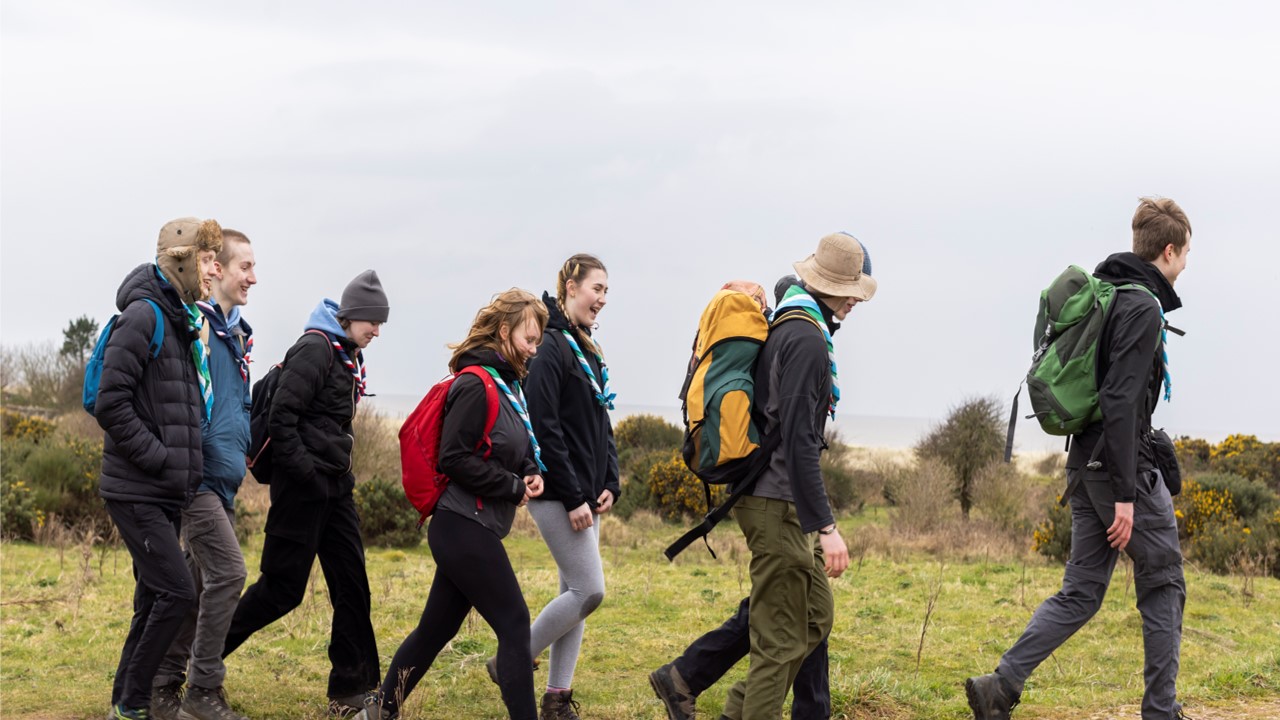 A group of eight young people carrying bags walk across a grass area.