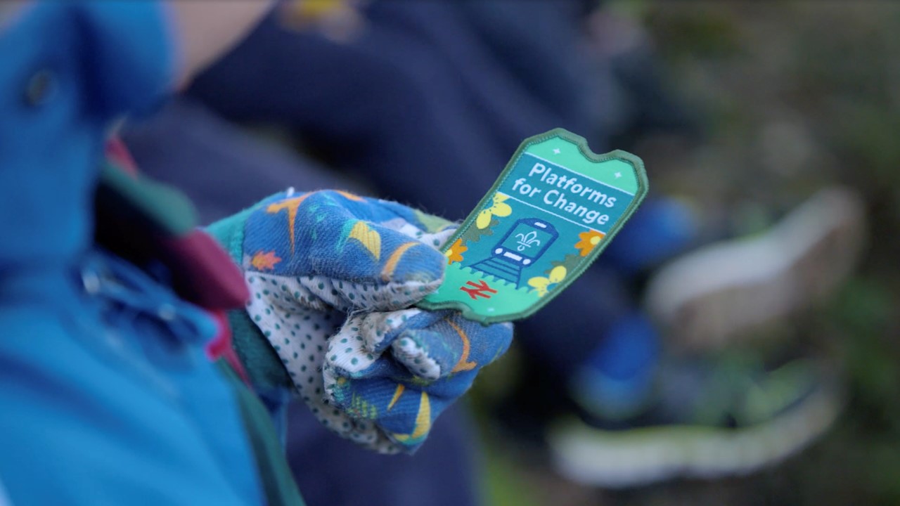 A young person wearing printed blue gloves holds the Platforms for Change badge.