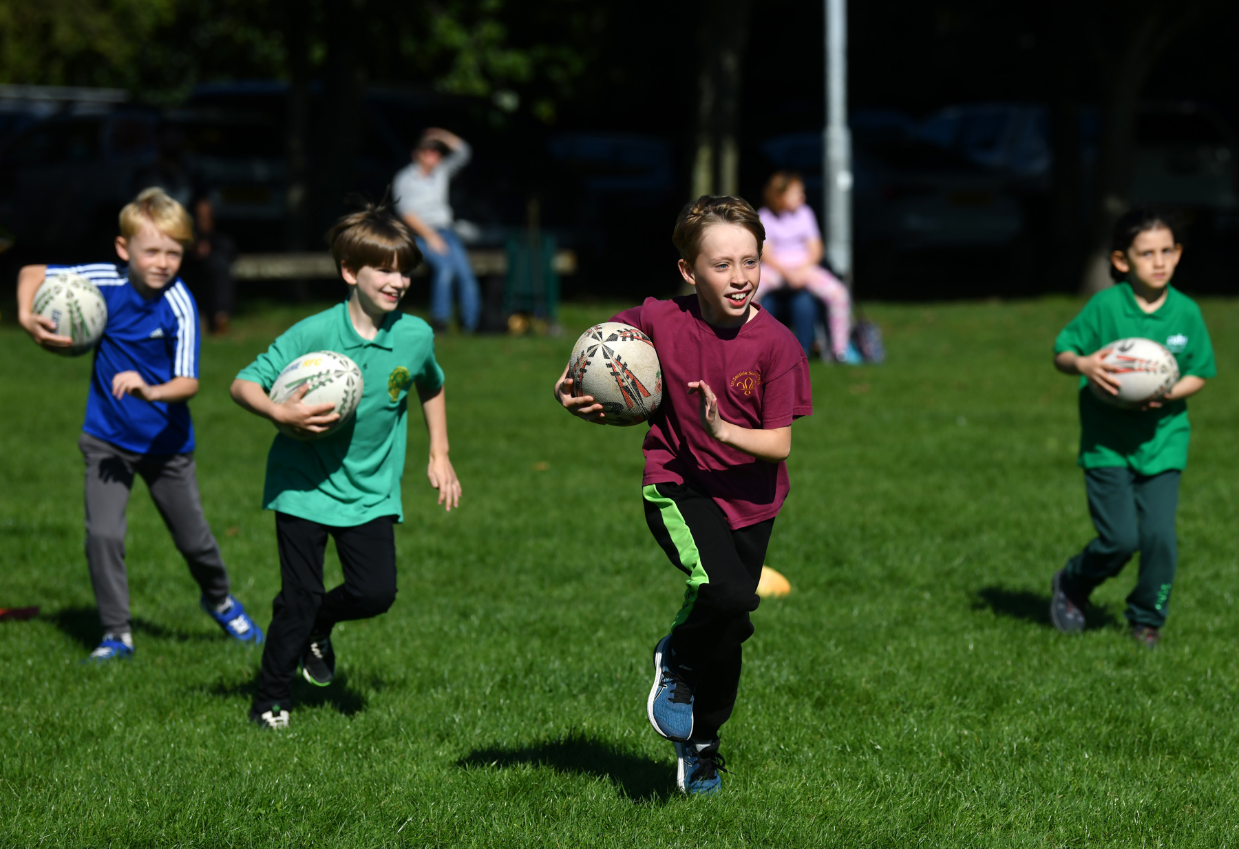 The image shows four Scouts on a grass playing field, each holding a rugby ball in their hands. They're each running forwards and they're wearing sports clothing.