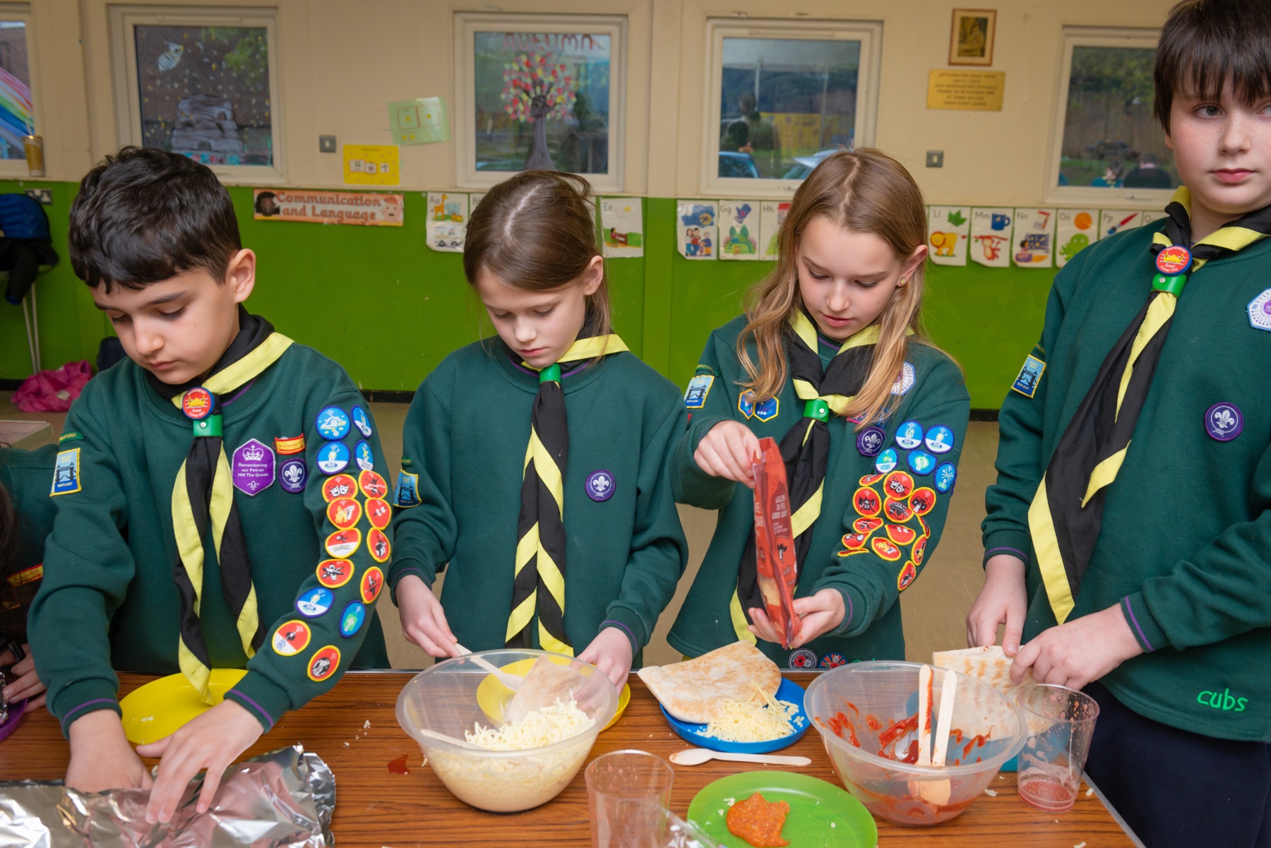 Four Cubs are stood behind a brown table with mixing bowls, plates, grated cheese and pitta breads in front of them. They're all in uniform and neckers, looking down at their plates. They're busy making pitta pockets.