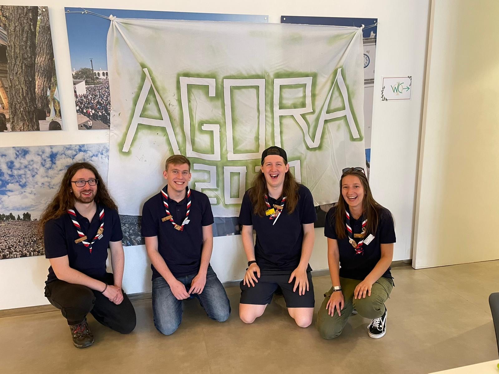 Four members of the UK Rep Pool are crouching down on their knees in front of a banner that reads 'Agora.' They're all wearing navy t-shirts and navy, red and white neckers, and smiling at the camera. 